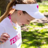 Girl turned to side wearing white visor with pink heart on it and white t shirt with tennis girl written on it from Love Set Match tennis wear
