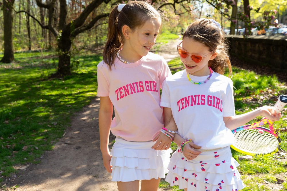 Two girls in cute tennis clothes from Love Set Match tennis wear. One is wearing a pink tennis tee and one is wearing a white tennis tee. Both are wearing cute tennis skirts with ruffles. 